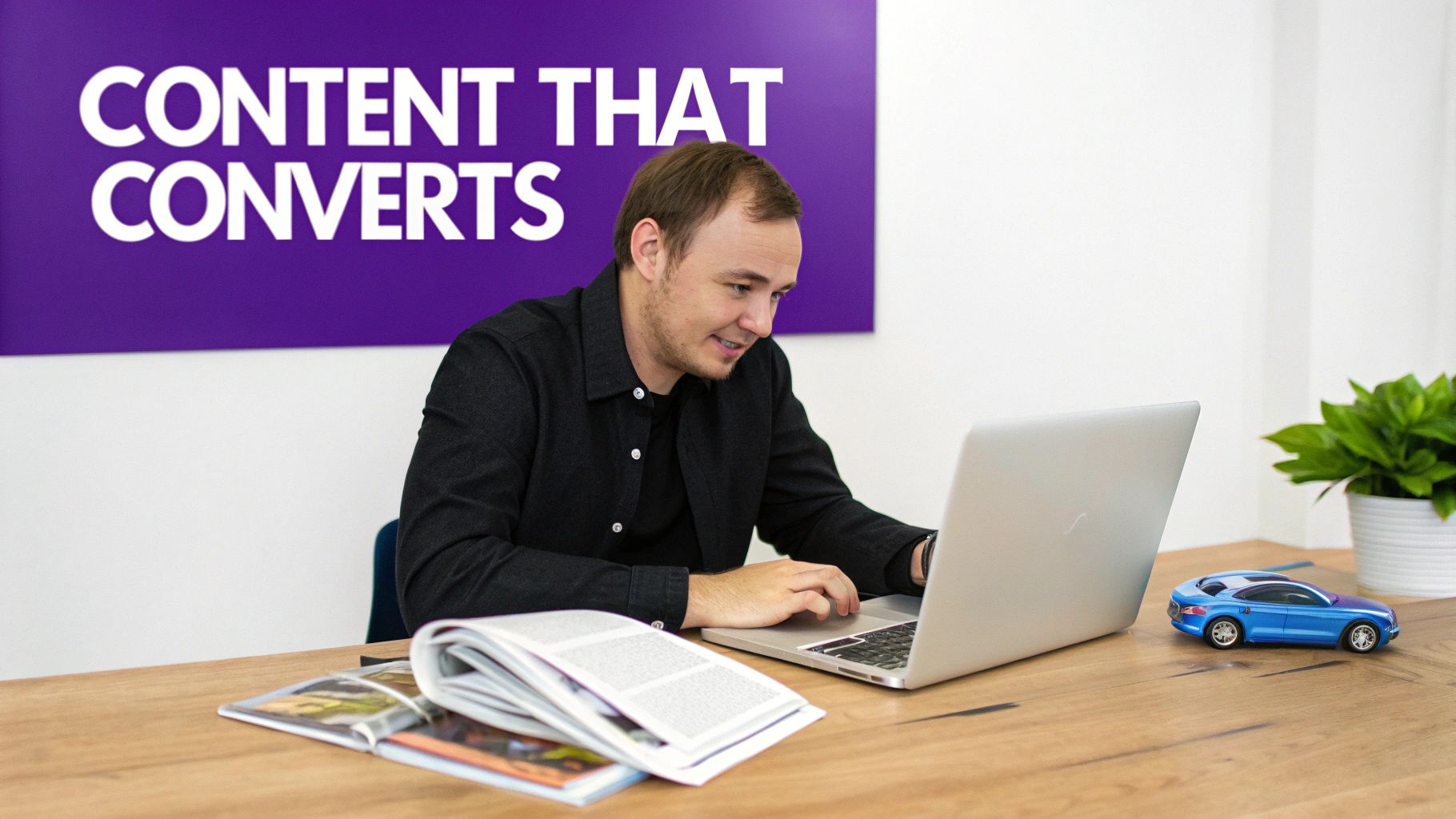 A man smiles while working on a laptop at a wooden desk, beside a toy car and plant.